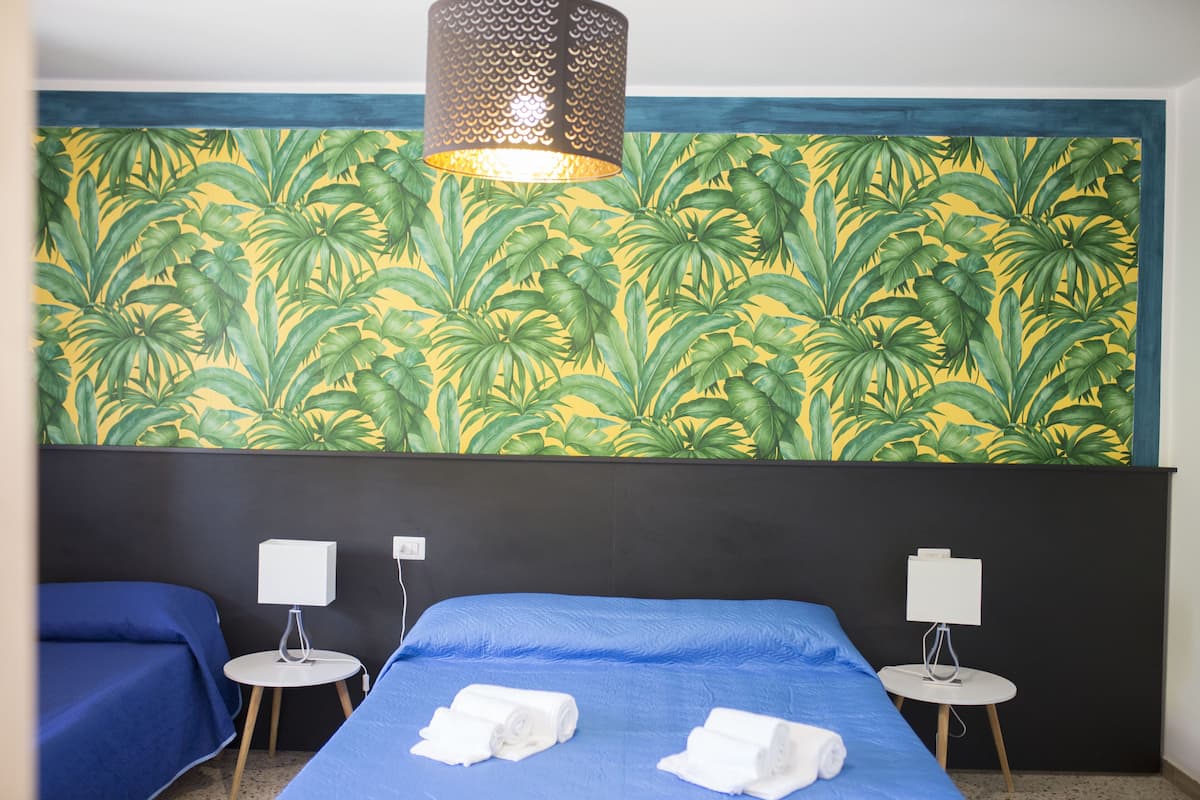 Habitación de hotel con cama, dos mesitas de noche, una lámpara y un papel tapiz con estampado de hojas tropicales.