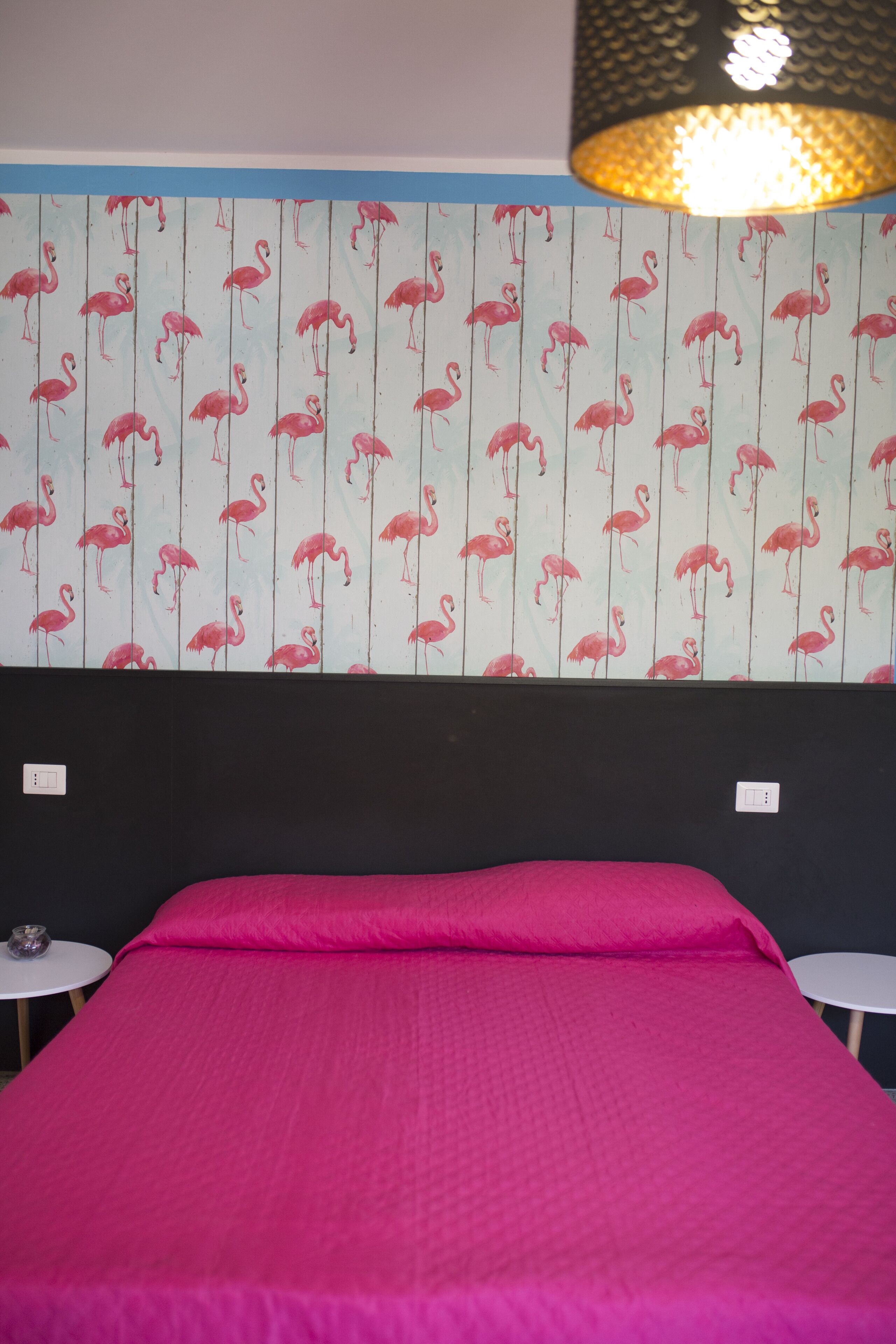 Een kamer met een bed, twee kleine tafels, een muur met een flamingo motief, en een hanglamp.