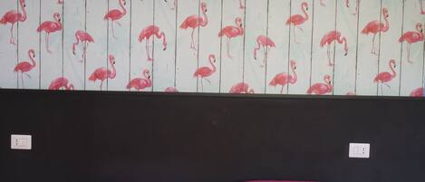Een kamer met een bed, twee kleine tafels, een muur met een flamingo motief, en een hanglamp.