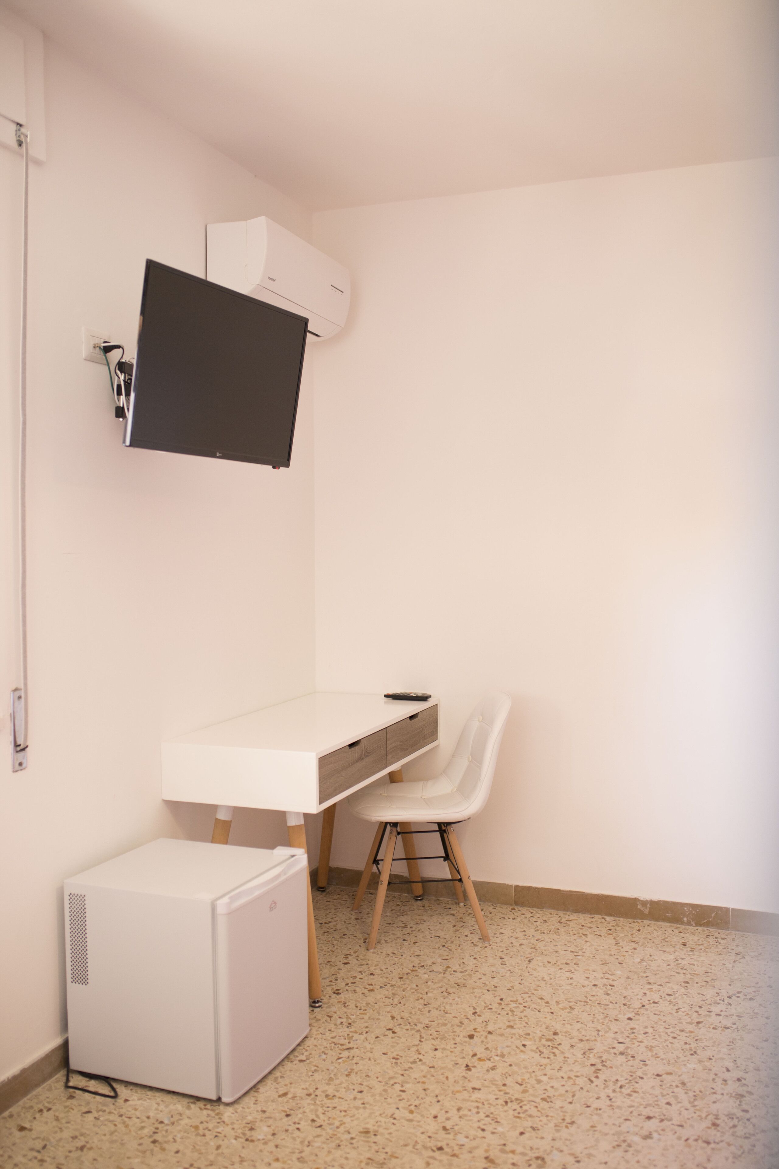 Une petite pièce propre, avec un bureau blanc, une chaise, un petit réfrigérateur blanc et un climatiseur fixé au mur.