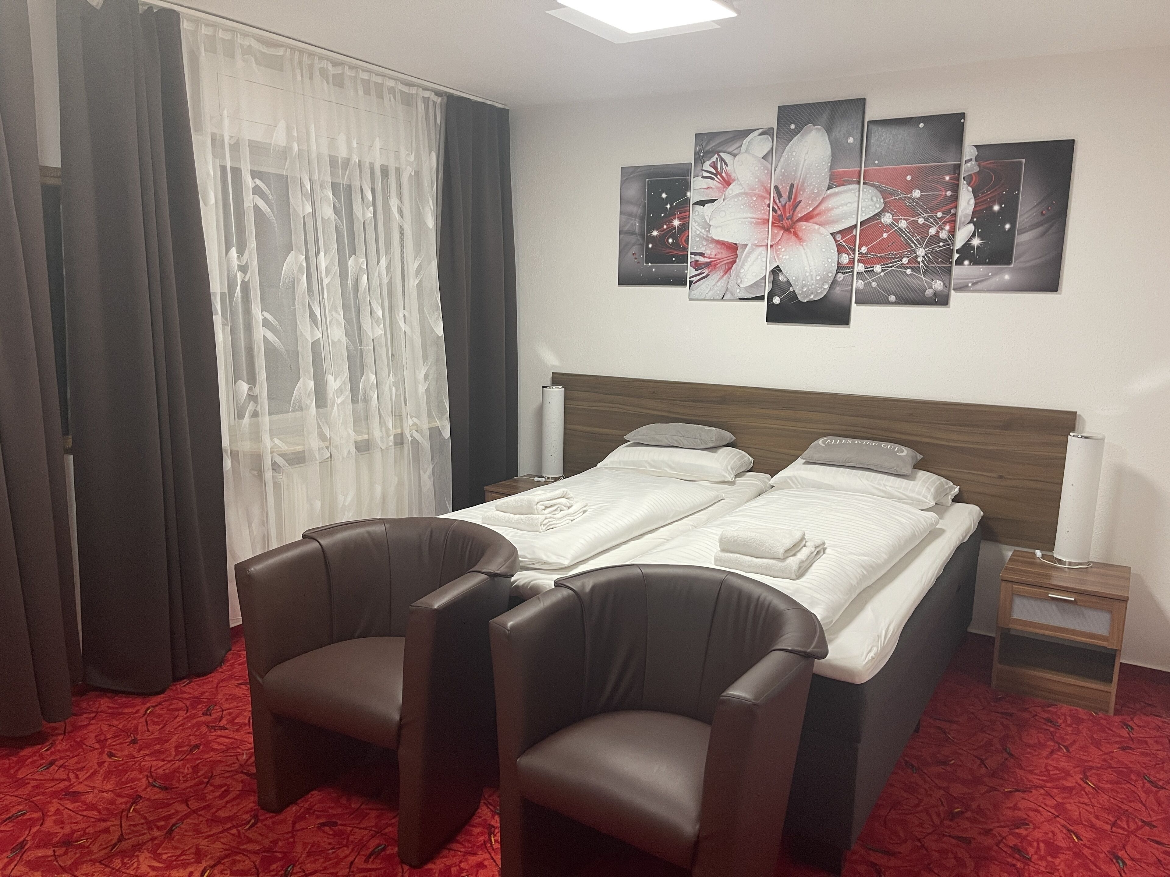 Habitación de hotel con dos camas, un ventanal con cortinas translúcidas, una alfombra roja y una pared decorada con tres cuadros.