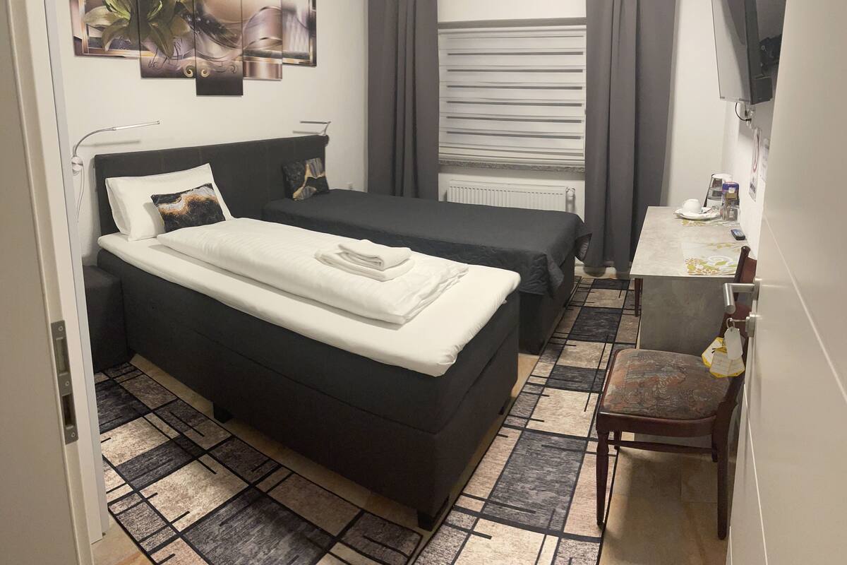 A hotel room with two beds, a desk, a chair, a radiator, and a window with curtains.
