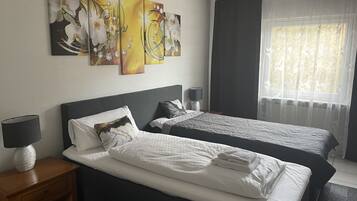 A hotel room with two beds, a nightstand with a lamp, and a window with sheer curtains.