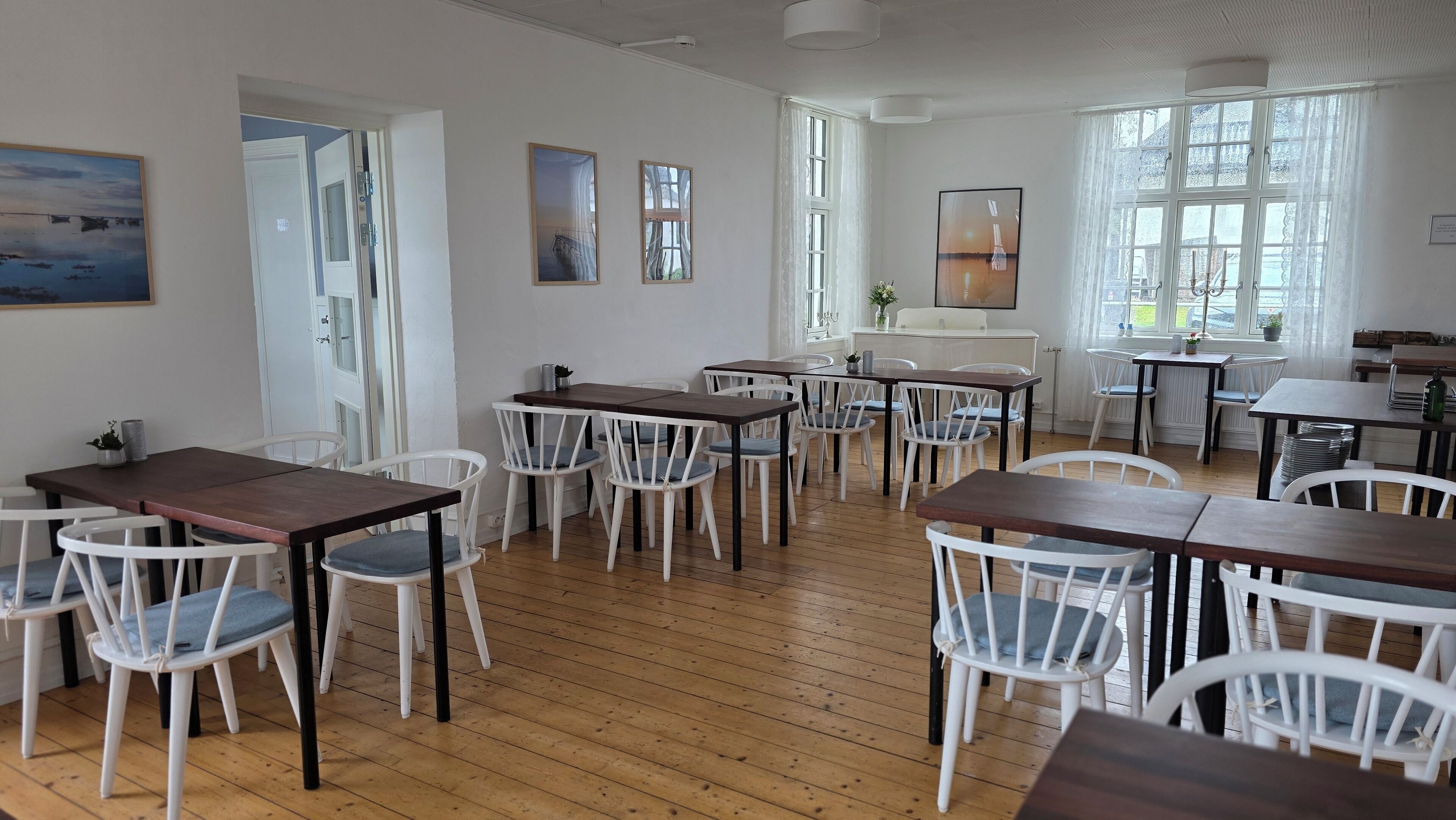 Un comedor con mesas de madera y sillas blancas, ventanas amplias con cortinas y obras de arte en las paredes.