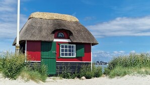 A small, thatched-roof building with a red and green facade, situated on a sandy beach with vegetation in the foreground and a clear sky in the background. - Hotel Udsigten Marstal (Marstal)