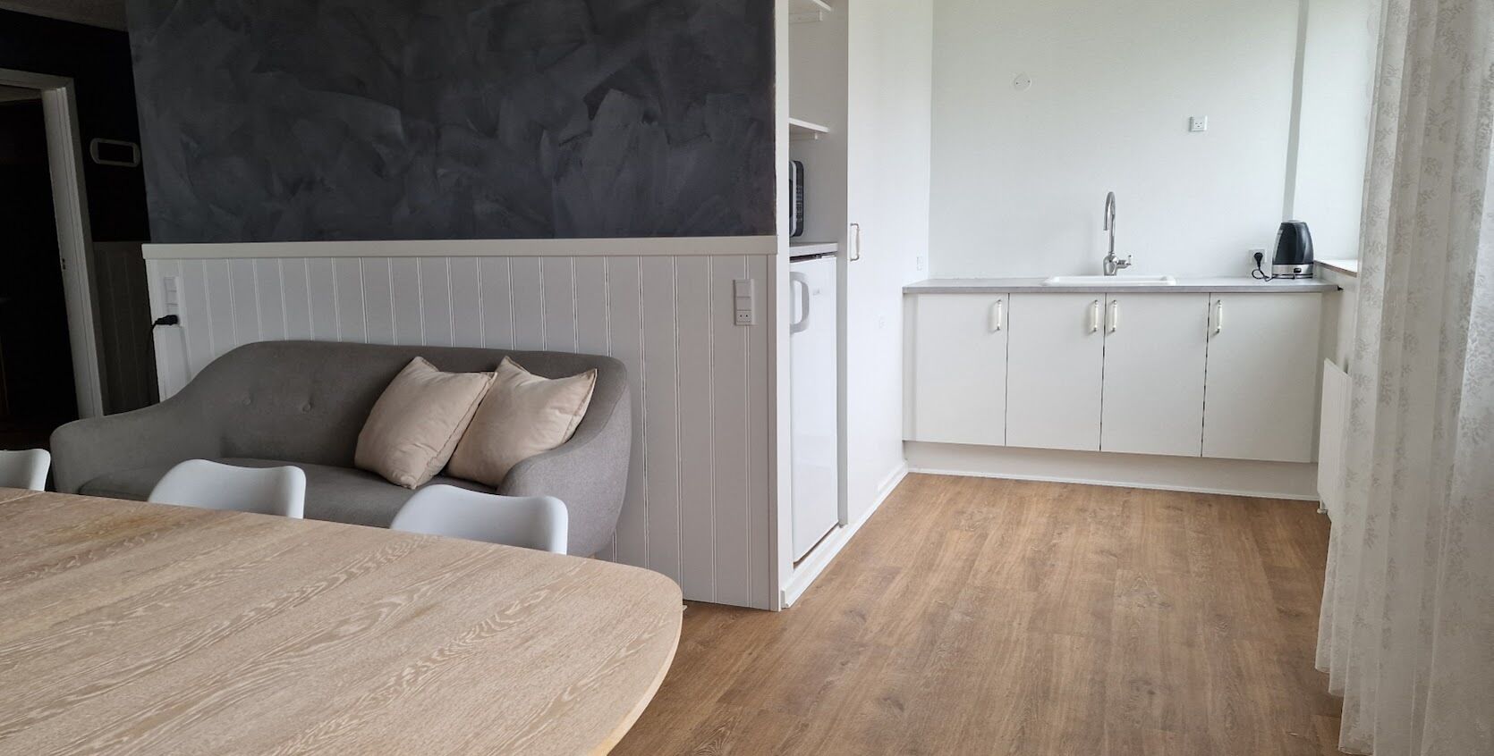 A modern room with a grey sofa, a wooden dining table, a white kitchenette, and a wooden floor.