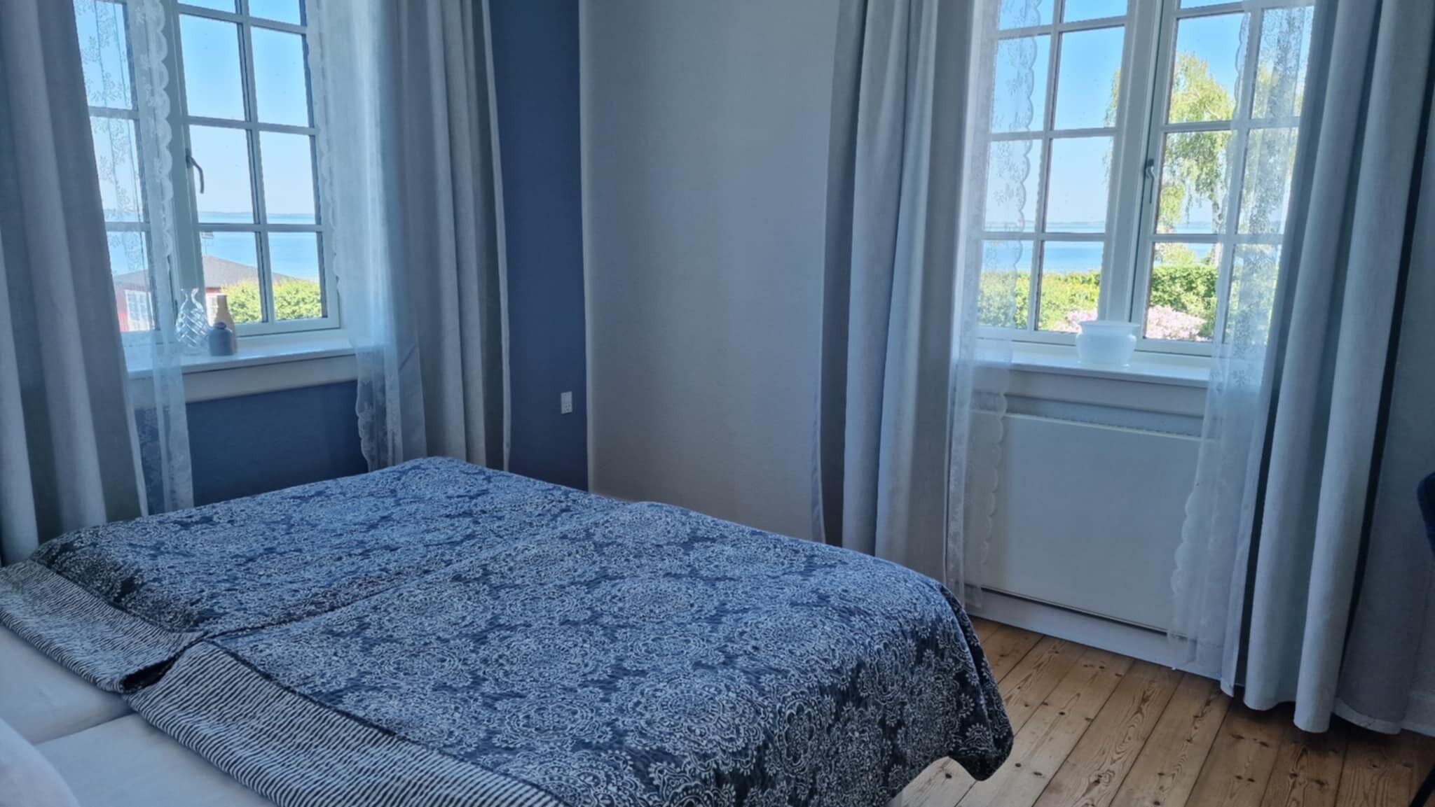 A room with a bed, two windows with curtains, a view of greenery outside, and a wooden floor.