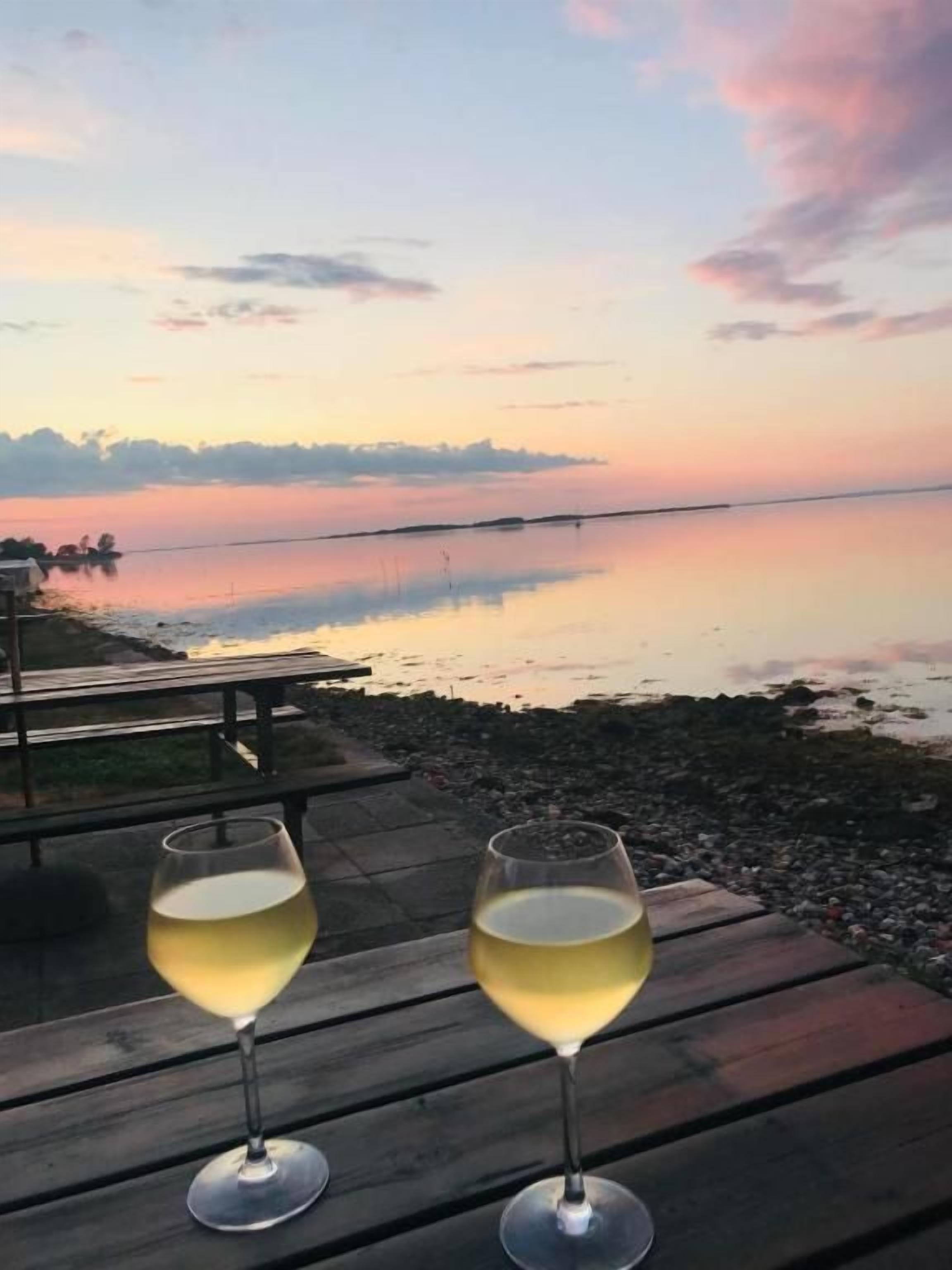 Deux verres de vin blanc sur une table en bois, avec vue sur une étendue d’eau calme et un coucher de soleil en arrière-plan.