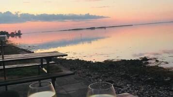 Deux verres de vin blanc sur une table en bois, avec vue sur une étendue d’eau calme et un coucher de soleil en arrière-plan.