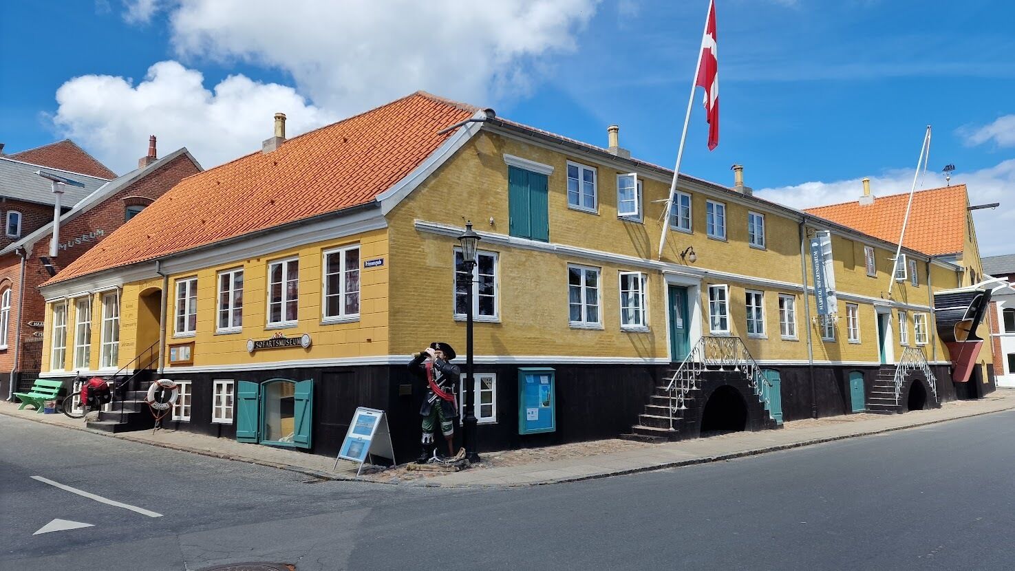Ein gelbes Gebäude mit rotem Ziegeldach, einer Fahnenstange mit einer Flagge und eine Person in Uniform vor dem Haus.