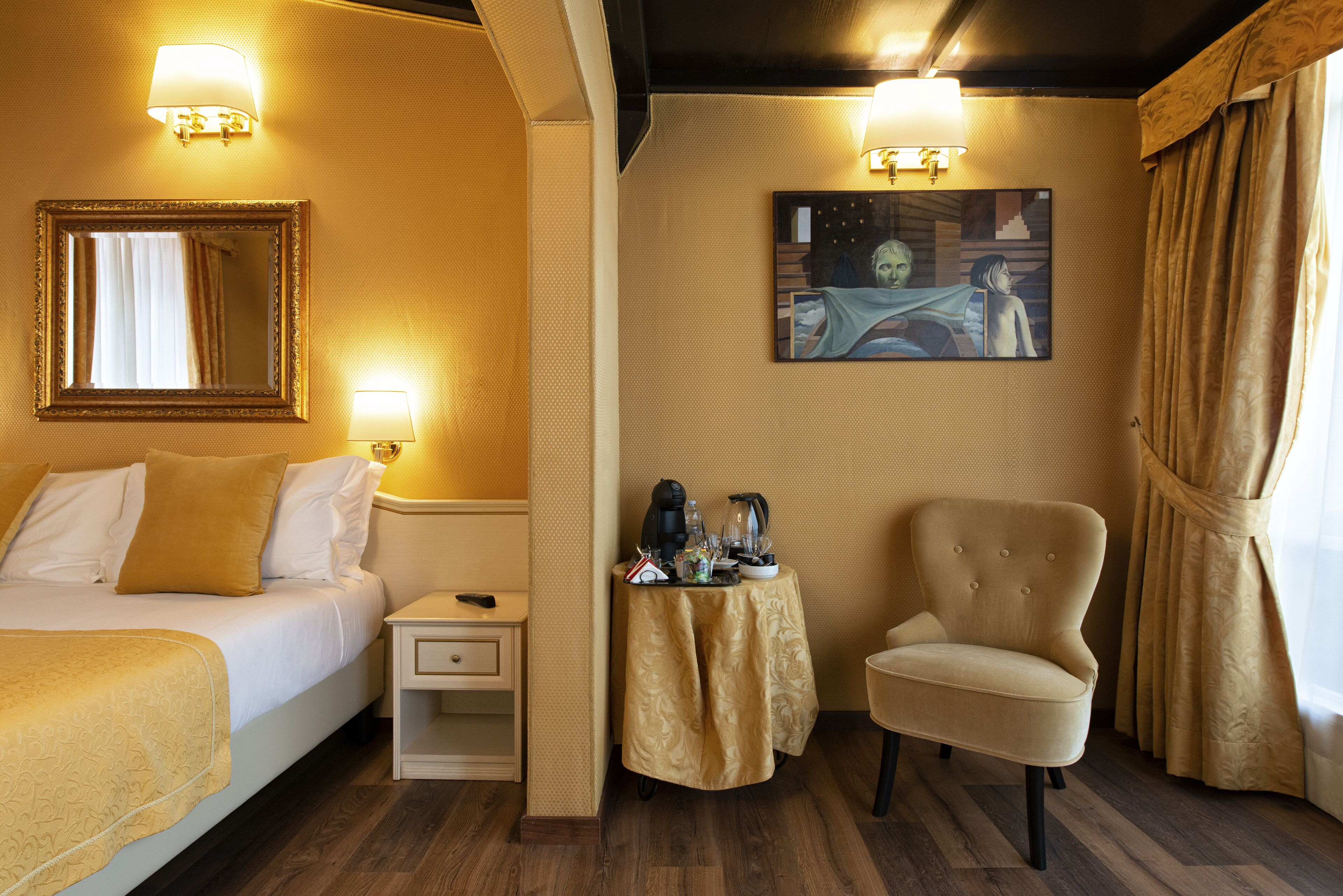 A hotel room with a bed, a chair, a nightstand, a table with a lamp, and a painting on the wall.