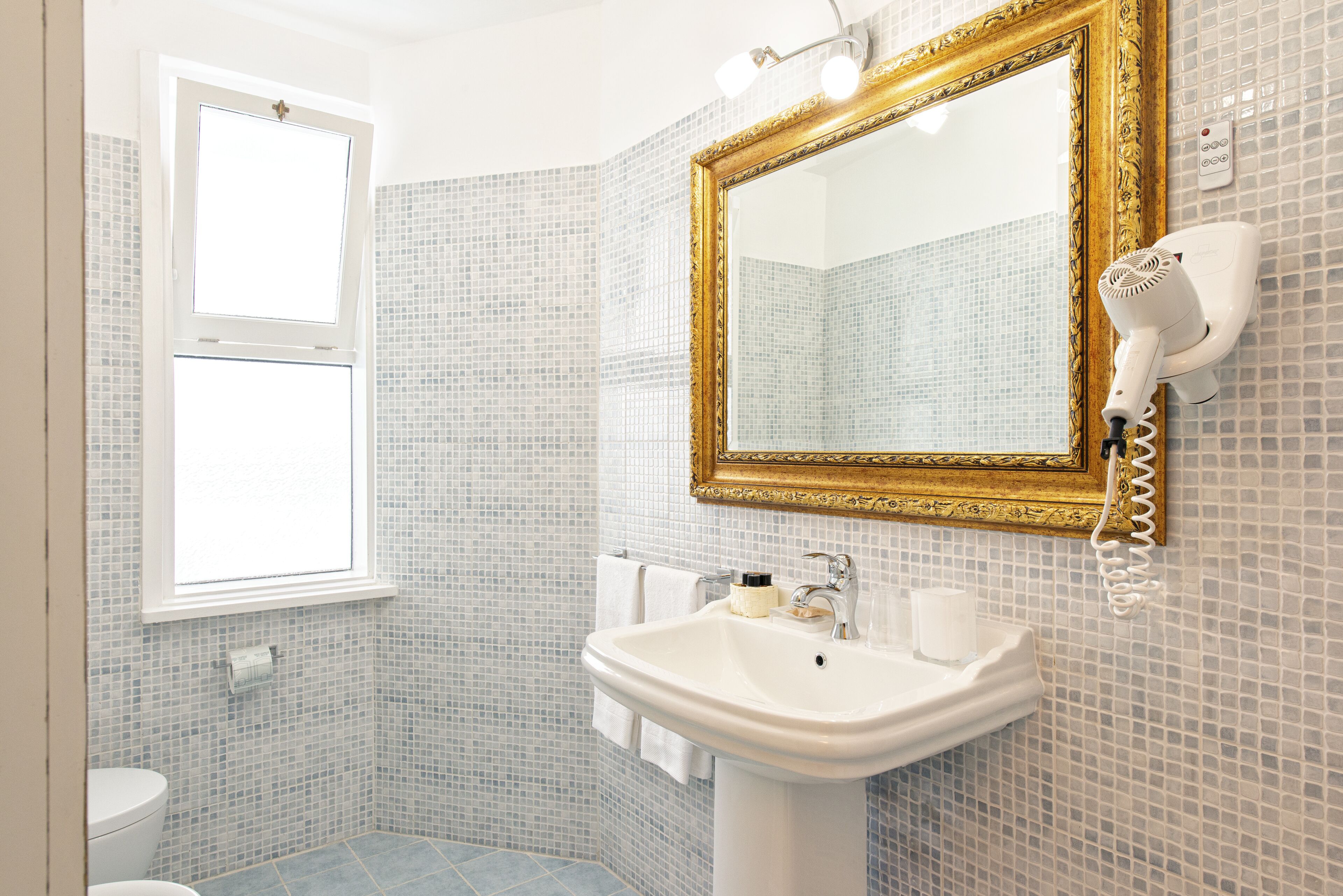 A bathroom with a white sink, a mirror with a gold frame, a hairdryer, and a window.