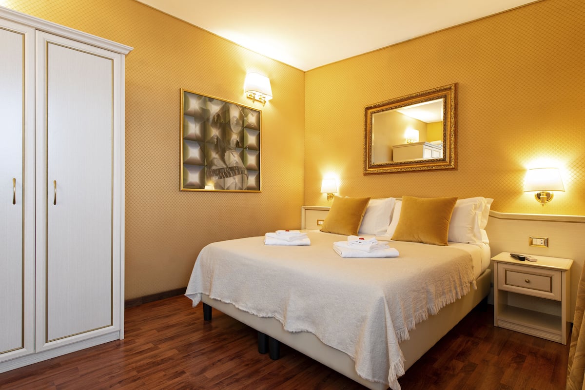 A hotel room with a large bed, a wardrobe, a painting, a mirror, and bedside lamps.