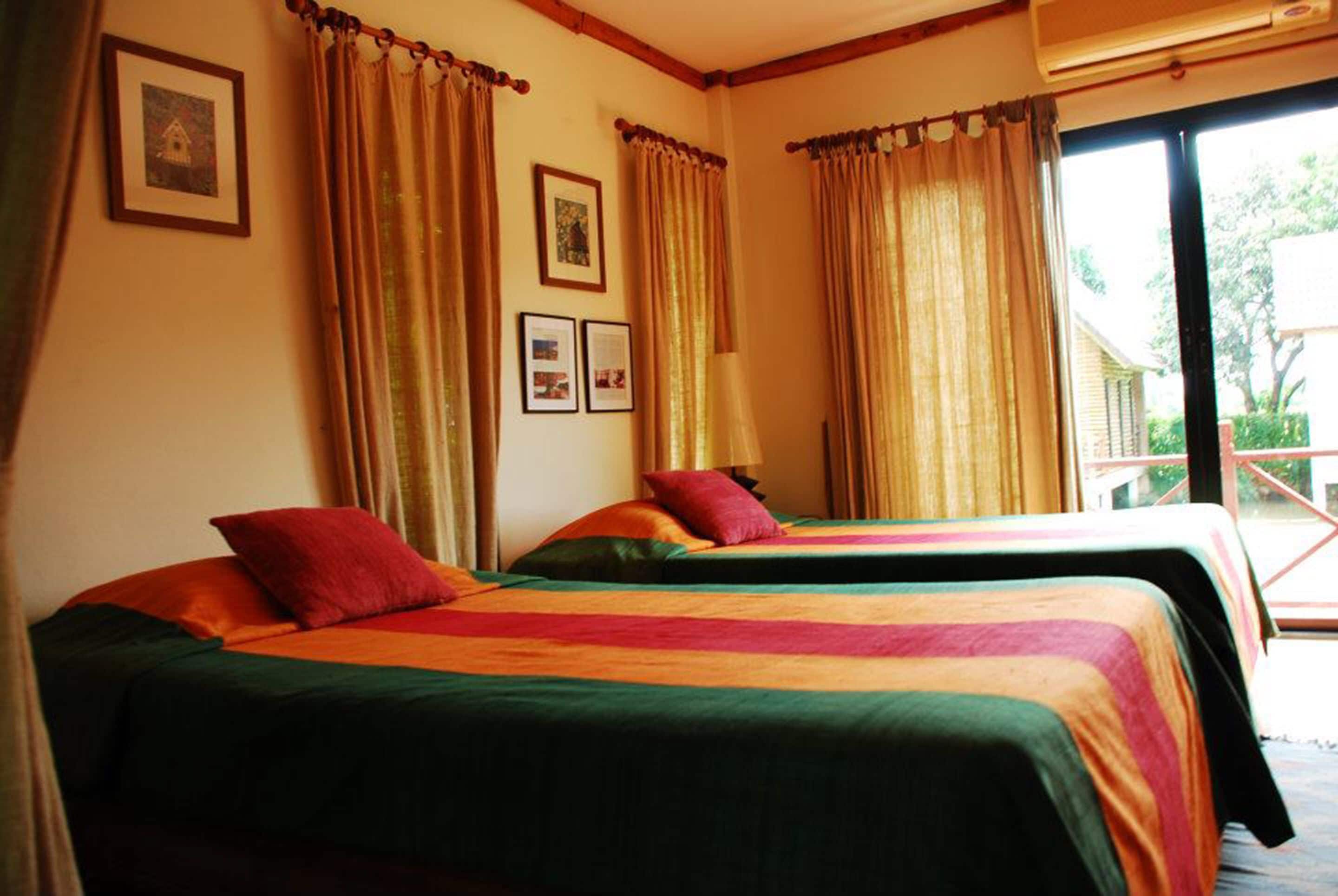Une chambre avec deux lits, chacun agrémenté de draps et d’oreillers aux couleurs vives, une fenêtre avec des rideaux et un climatiseur fixé au mur.