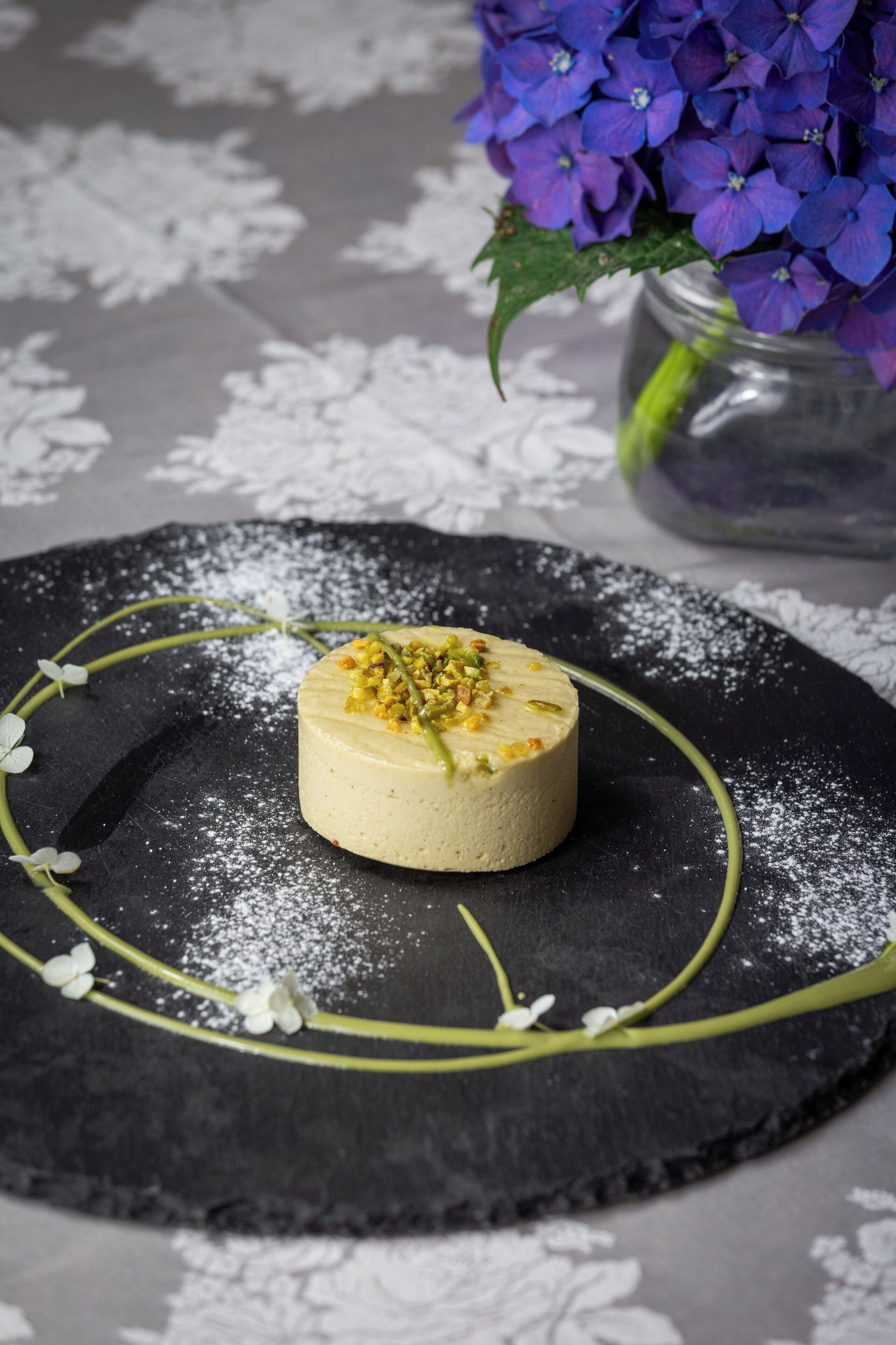 A dessert on a black plate with powdered sugar and green garnish, surrounded by white flowers, on a table with a lace tablecloth.