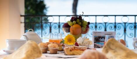 Desayuno con pasteles, un bol de fruta, una tetera y un vaso de jugo de naranja en un balcón con baranda de metal y vista al cielo.