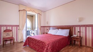 A bedroom with a wooden bed, a nightstand with a phone, a chair with a 'Holiday' pillow, and a balcony with a table and chairs. - Hotel Holiday (Bolsena)