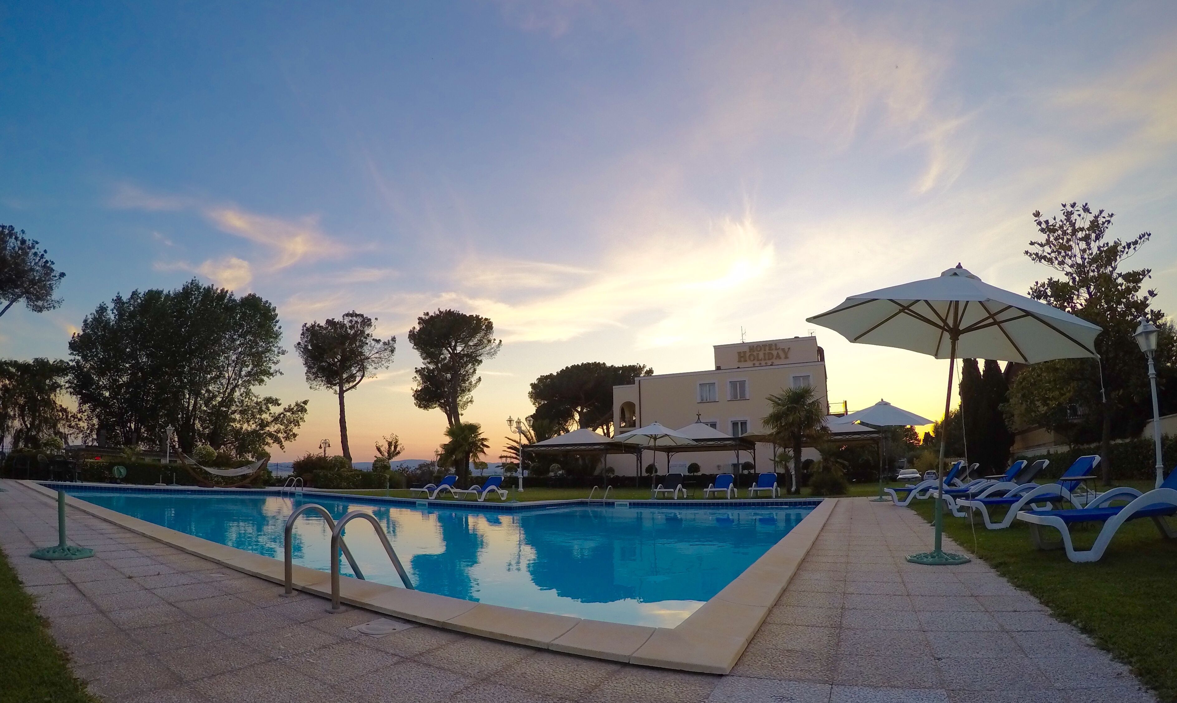 Ein Swimmingpool mit Liegestühlen und Sonnenschirmen, umgeben von Bäumen und einem Gebäude mit der Aufschrift 'HOTEL HOLIDAY'.