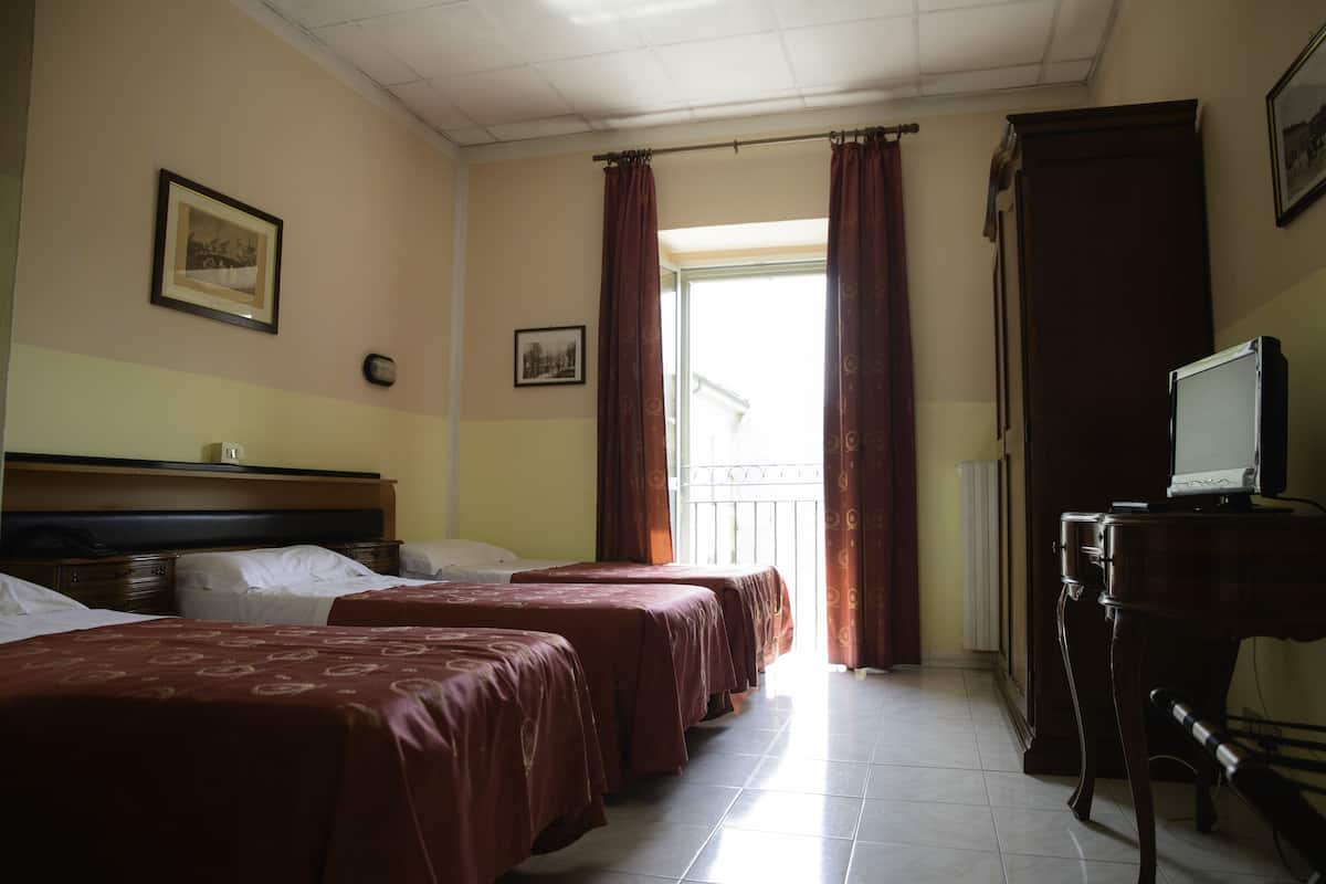 A hotel room with two beds, a desk with a computer, a radiator, and a window with curtains.