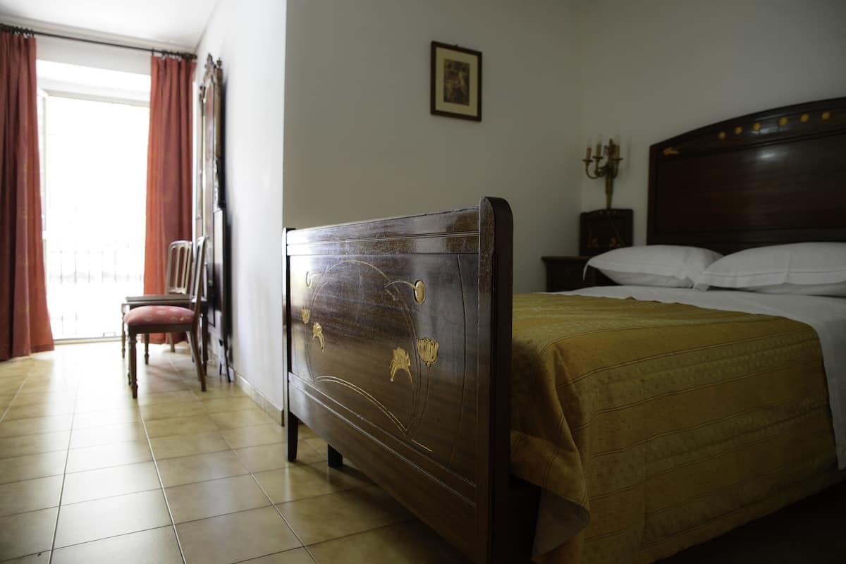 A hotel room with a wooden bed, a chair, a bedside table with a lamp, and a window with curtains.
