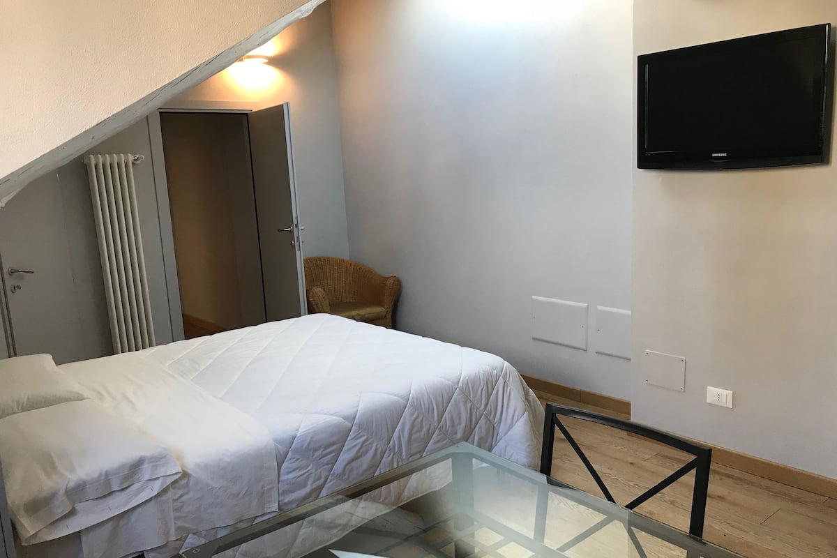 A bedroom with a bed, a glass table, a television mounted on the wall, and a wicker chair.