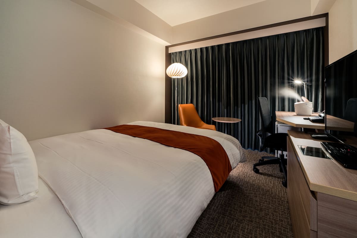 A hotel room with a bed, a desk, a chair, a lamp, and a television.