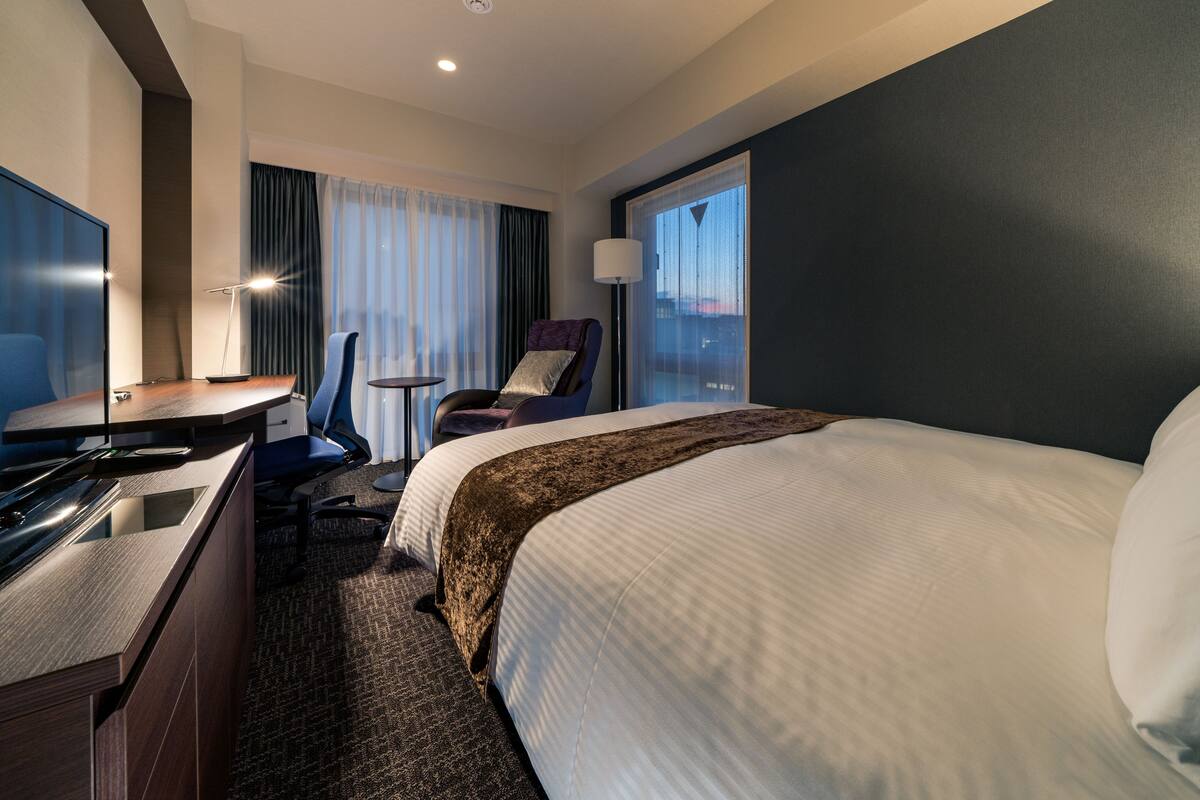 A hotel room with a bed, desk, chair, and a view of the city through the window.