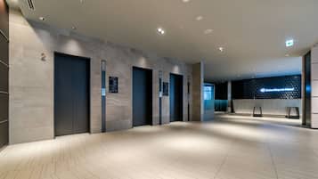 A spacious lobby with a modern design, featuring a herringbone-patterned floor, multiple elevators, and a reception area with a digital sign.