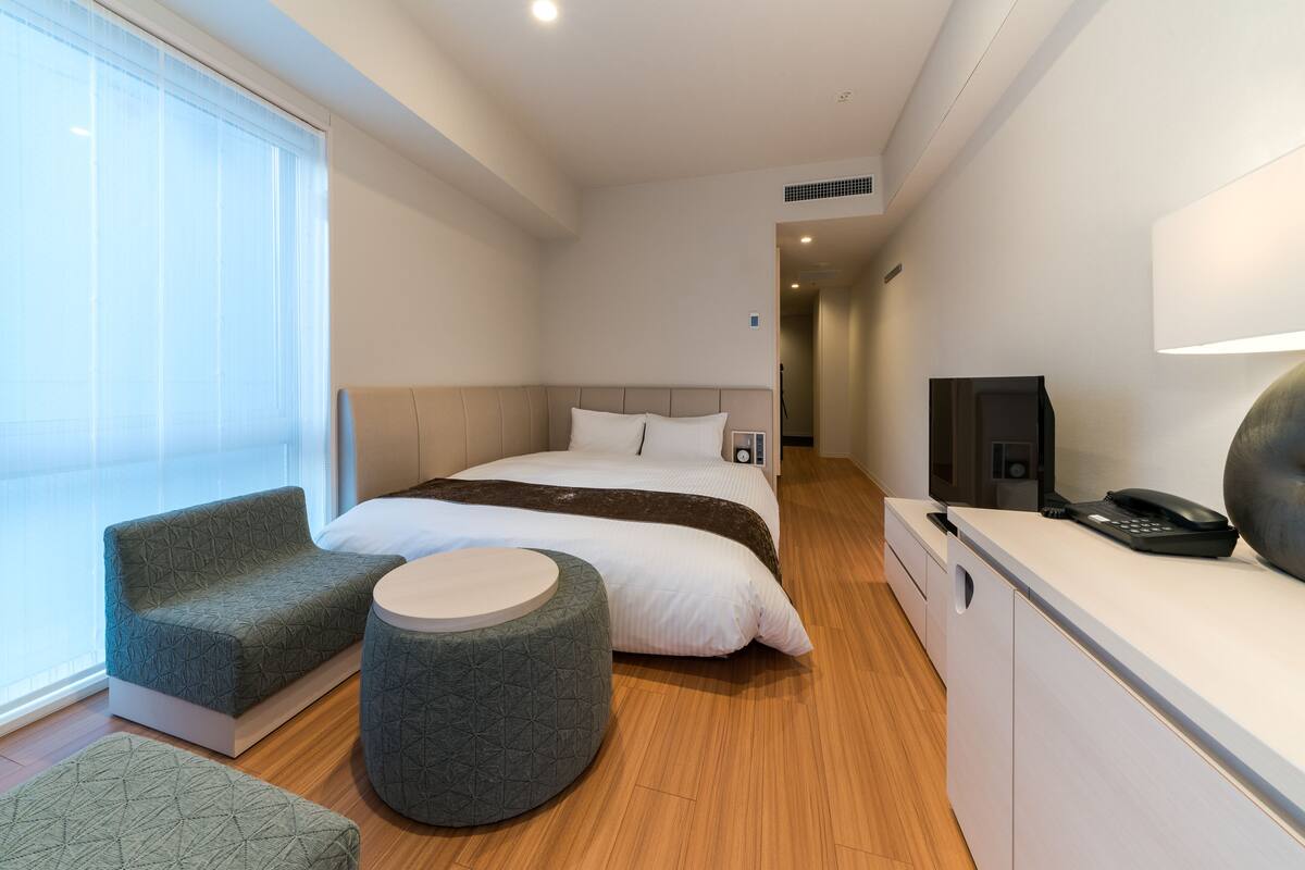 A modern hotel room with a bed, a sofa, a round ottoman, a desk with a phone, and a TV.