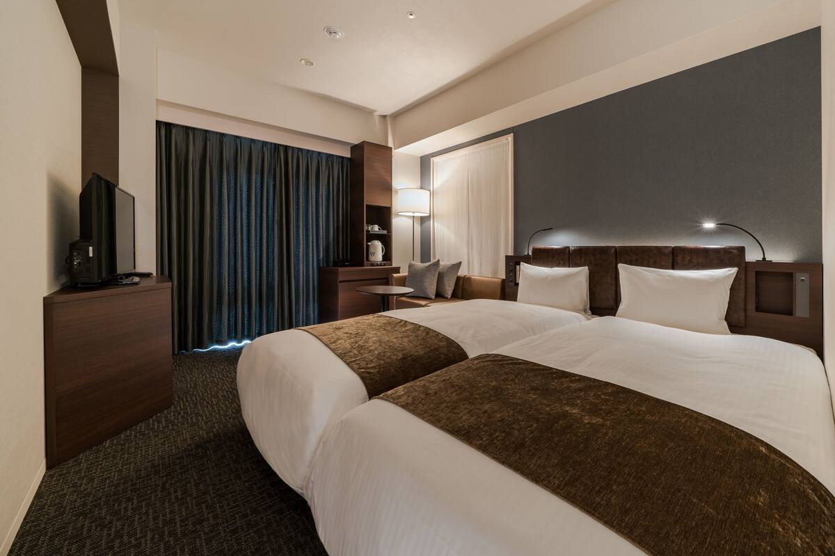 A hotel room with two beds, a desk, a TV, a lamp, and a large window with curtains.