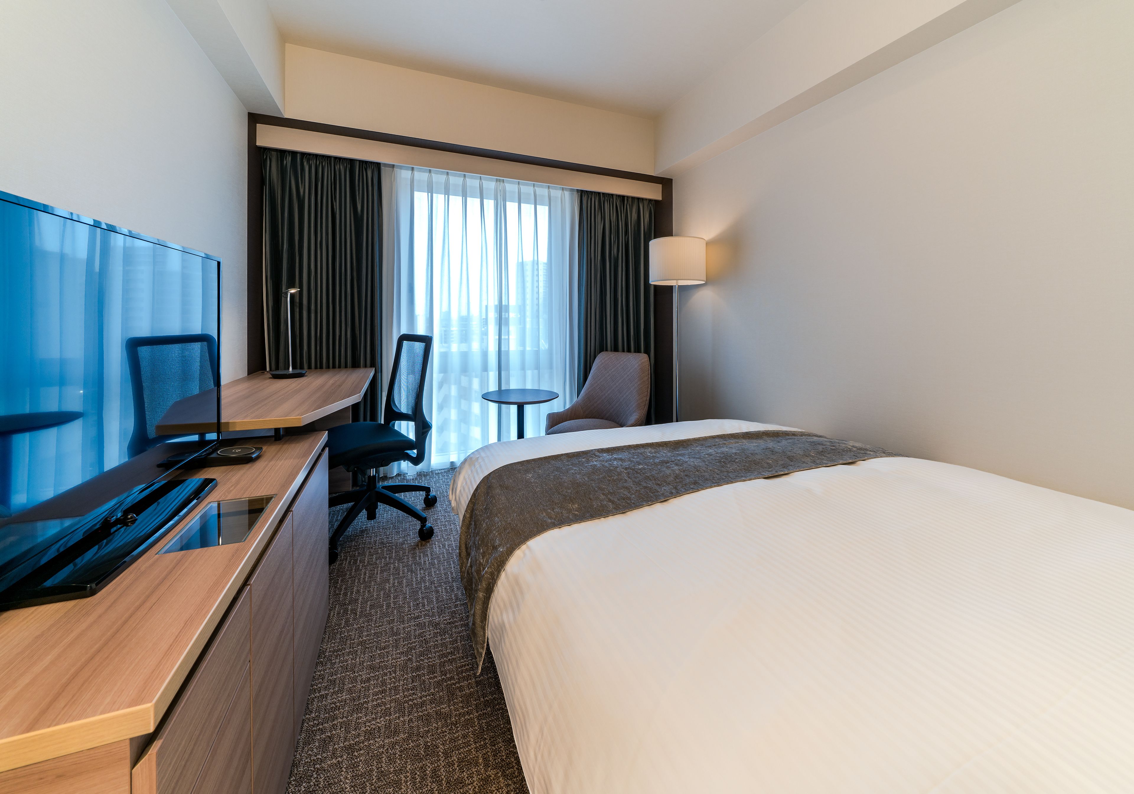 A hotel room with a bed, a desk with a computer, a chair, and a lamp.