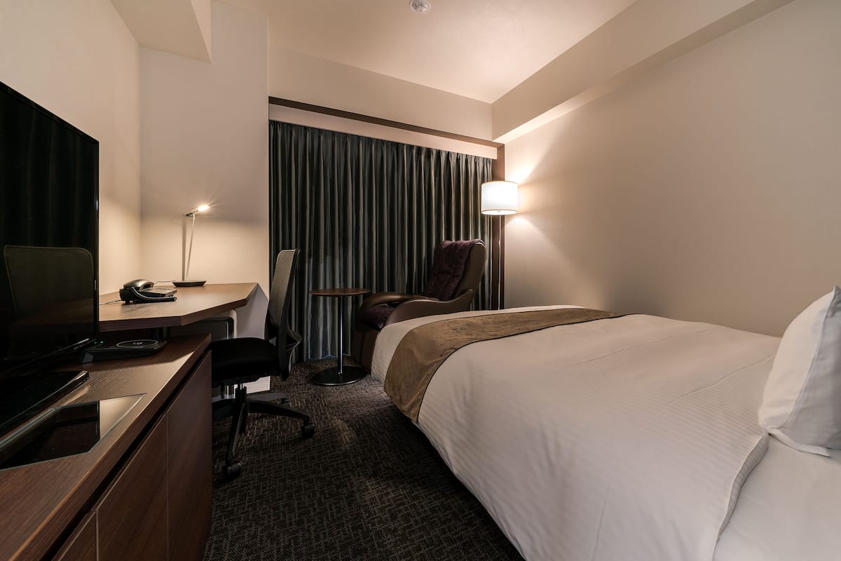 A hotel room with a bed, a desk with a computer, a chair, a TV, and a lamp.