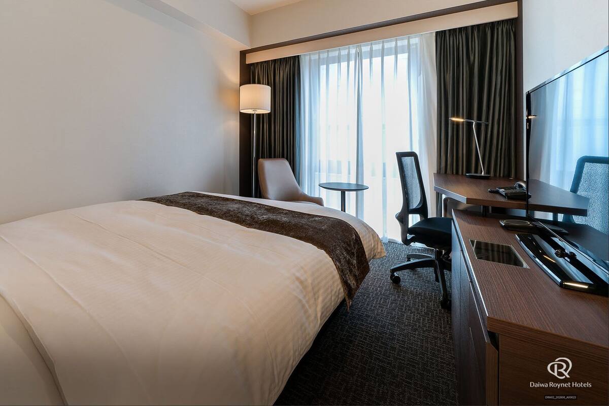 A hotel room with a bed, a desk with a computer, a chair, and a TV.