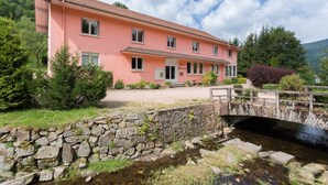 A pink building with a stone bridge over a stream, surrounded by greenery and hills. - Les Sources (Bussang)