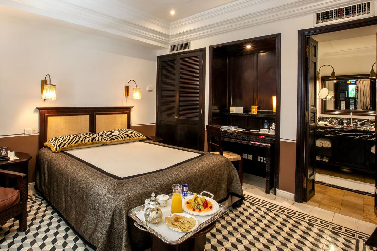 A hotel room with a bed, a desk, a chair, a tray with breakfast, and a bathroom visible through an open door.