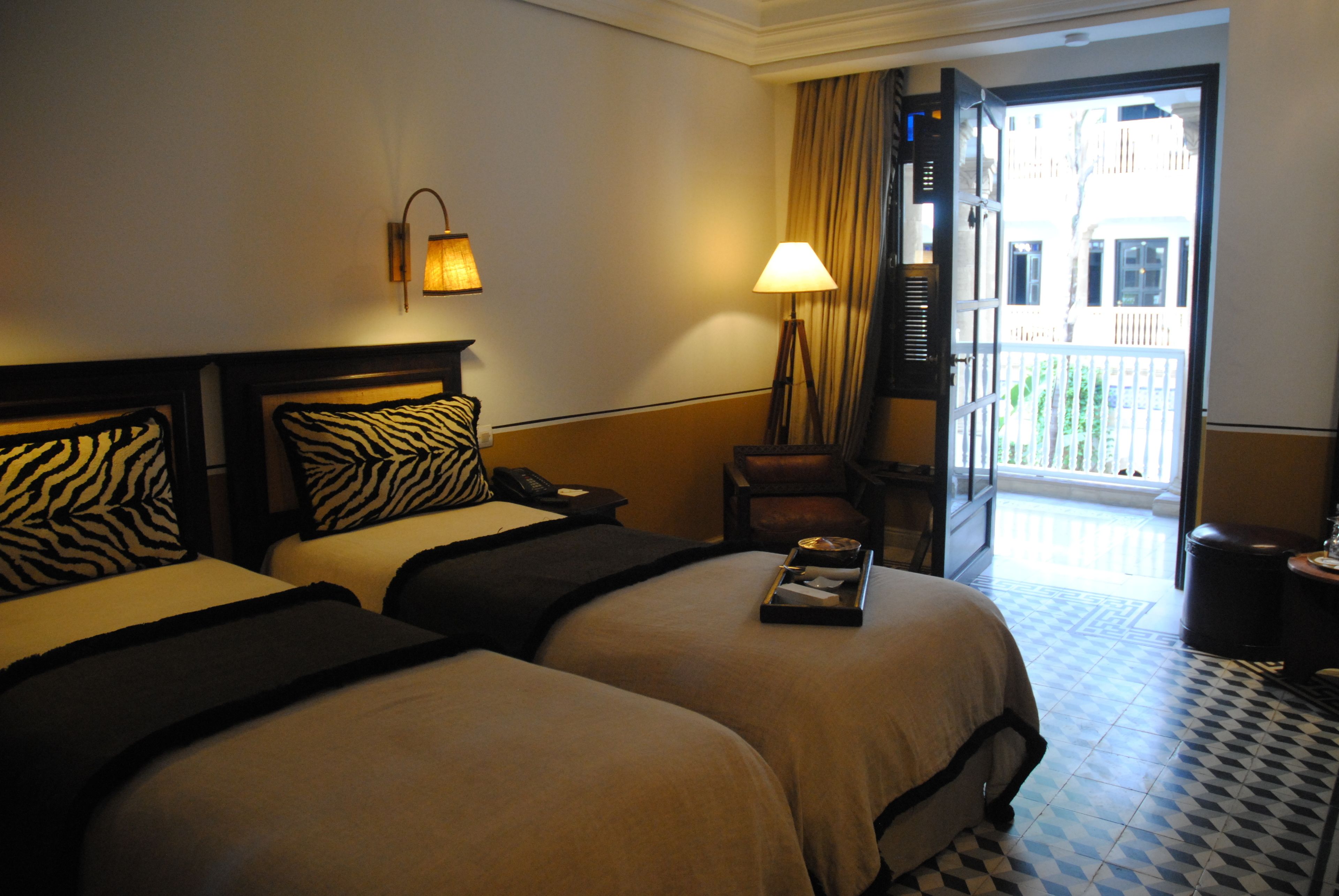 Habitación de hotel con dos camas, una mesita de noche, una lámpara, una silla y vistas al balcón exterior.