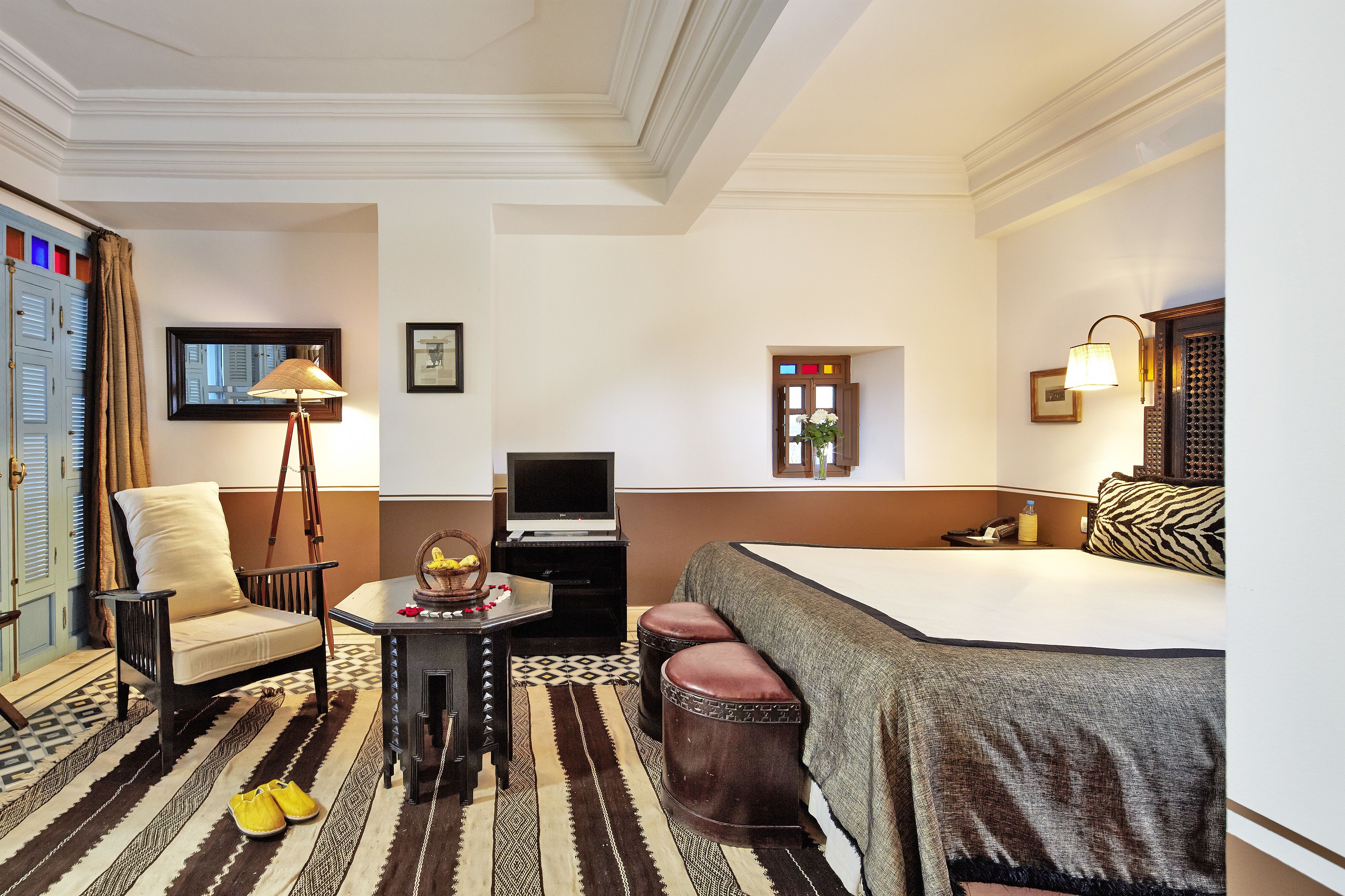 Habitación de hotel con cama, escritorio con lámpara, silla, pequeña mesa con un objeto decorativo y alfombra estampada.