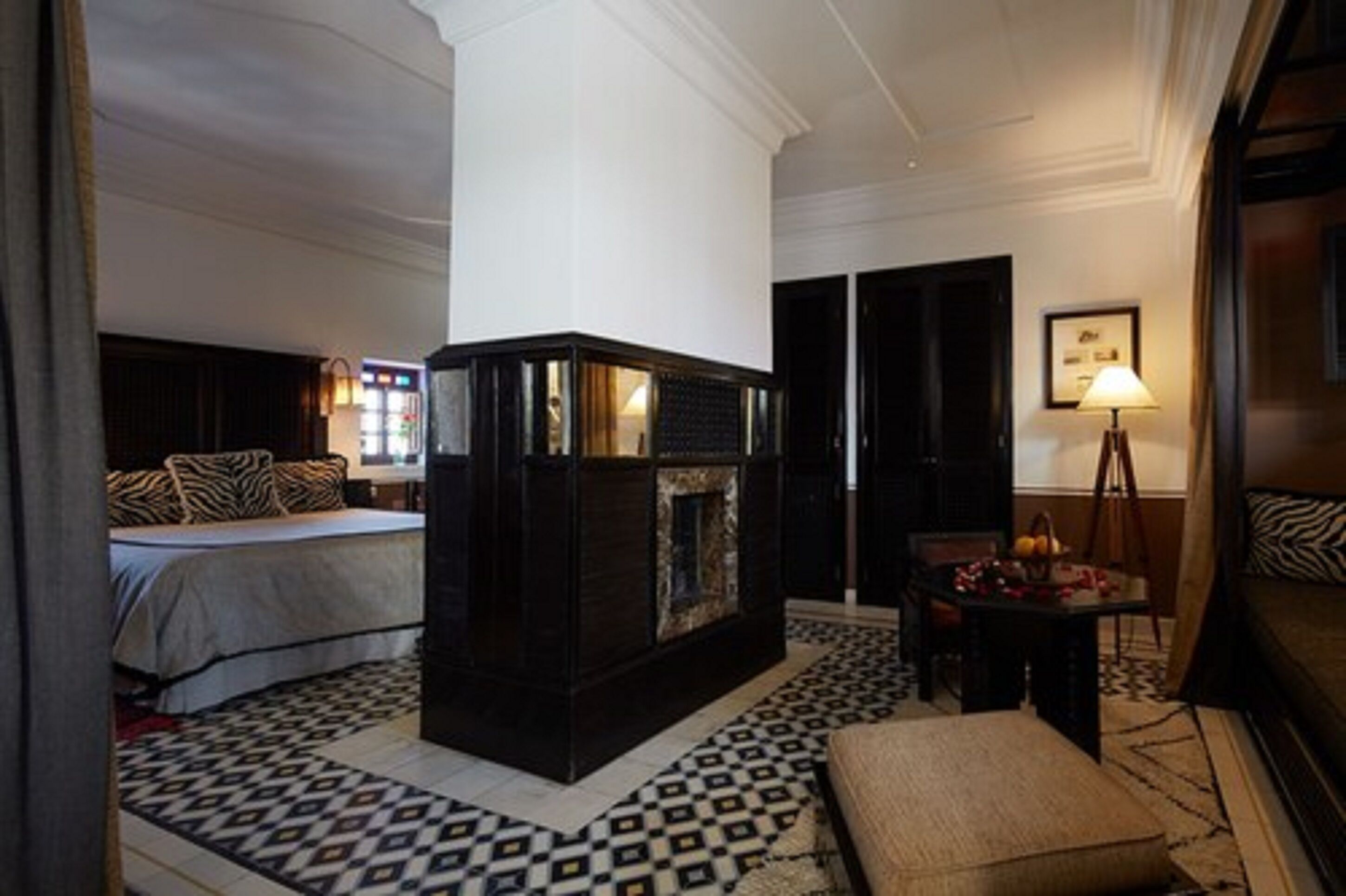 A hotel room with a large bed, a dark wooden fireplace, a patterned rug, and a small table with a lamp.