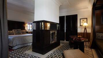 A hotel room with a large bed, a dark wooden fireplace, a patterned rug, and a small table with a lamp.