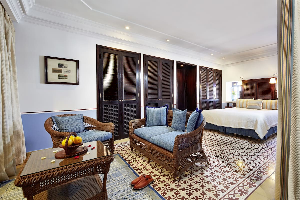 A hotel room with a large bed, two wicker sofas, a glass coffee table with fruit, and a patterned rug.