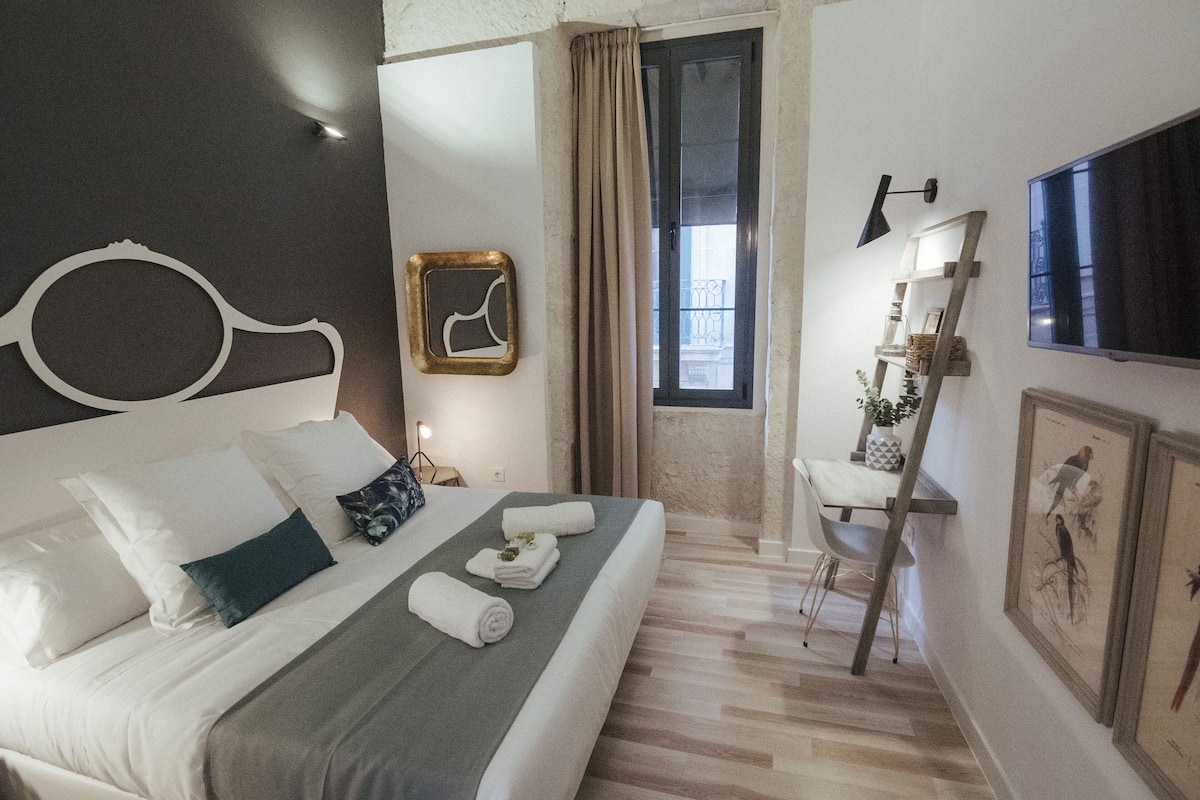 A neatly made bed with white and gray linens, a bedside table with a lamp, a window with curtains, and framed pictures on the wall.