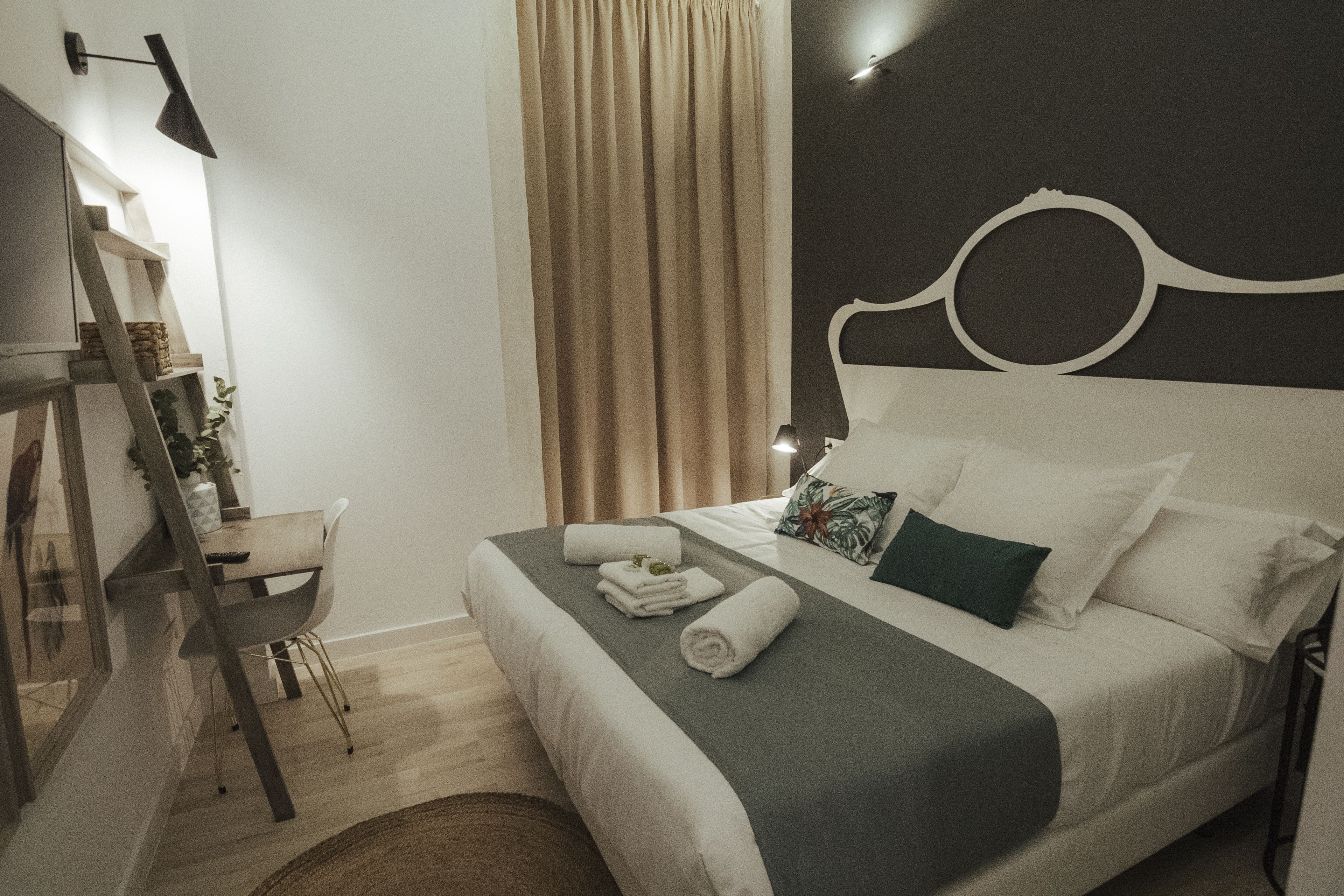 Una cama bien hecha con cubrecama gris y blanco, una silla de madera, un escritorio con lámpara y un ventanal con cortinas.