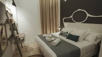 Una cama bien hecha con cubrecama gris y blanco, una silla de madera, un escritorio con lámpara y un ventanal con cortinas.