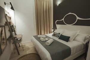 Uma cama bem arrumada com colcha cinza e branca, uma cadeira de madeira, uma escrivaninha com abajur e uma janela grande com cortinas.