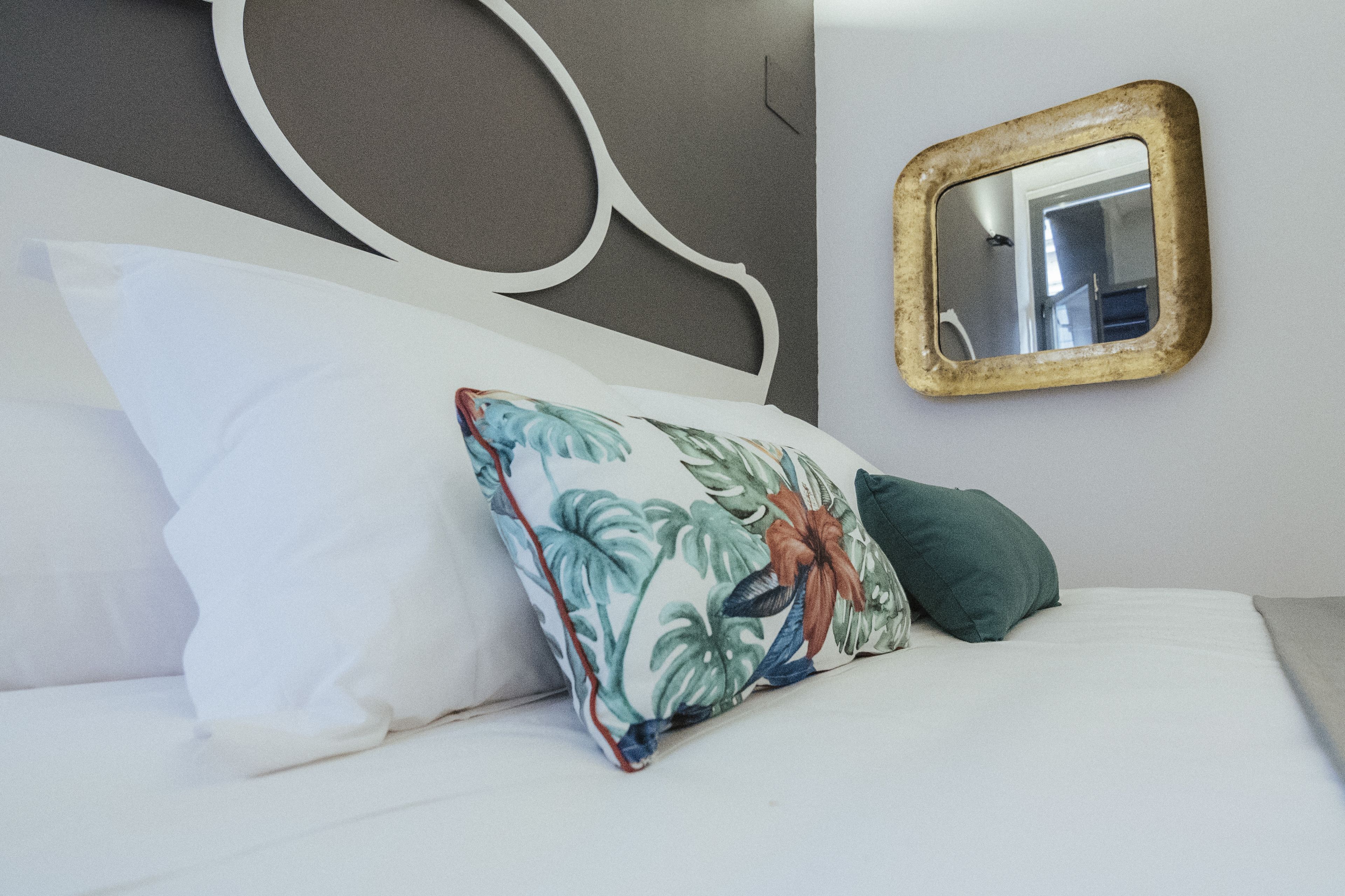 Una cama bien tendida con un almohadón floral y uno verde, un cabecero de madera y un espejo en la pared.
