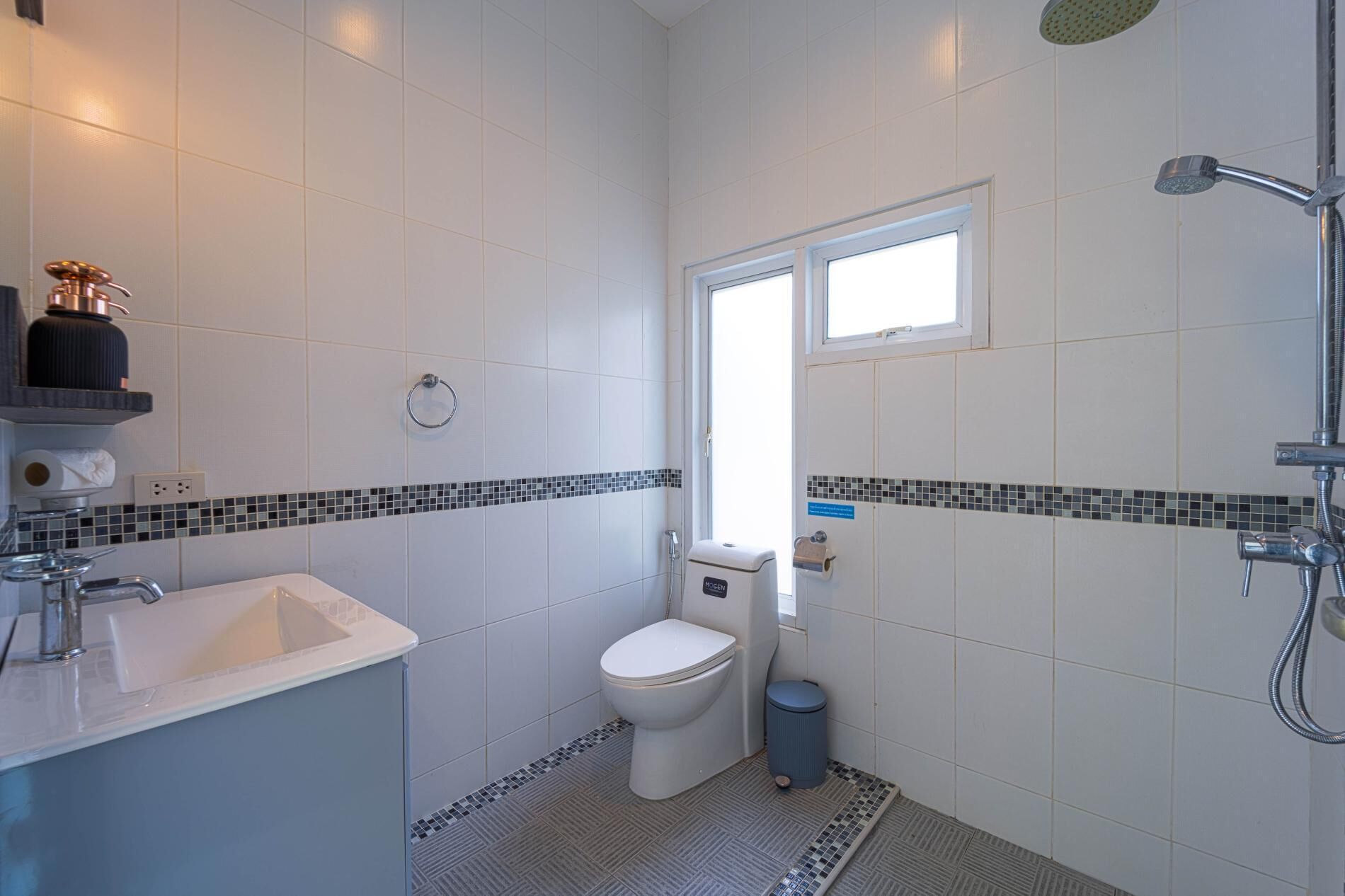 A small, modern bathroom with white tiles, a toilet, a sink, a shower, and a window.
