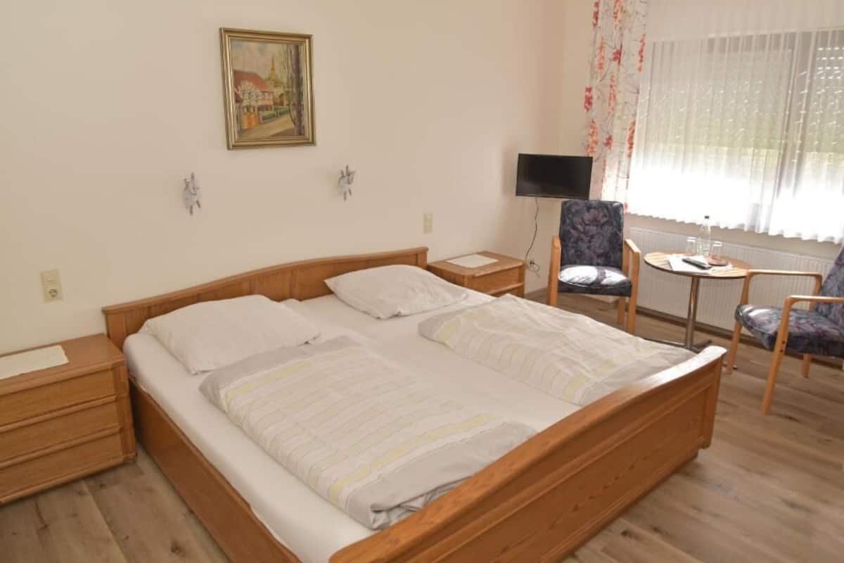 A bedroom with a wooden bed, a desk, a chair, a TV, and a window with curtains.