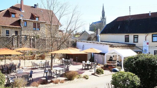 An outdoor cafe with tables and chairs, a white building with a sign, and a church in the background. - Weinhaus Hehner Kiltz (Waldböckelheim)