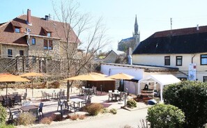 An outdoor cafe with tables and chairs, a white building with a sign, and a church in the background. - Weinhaus Hehner Kiltz  (Waldböckelheim)