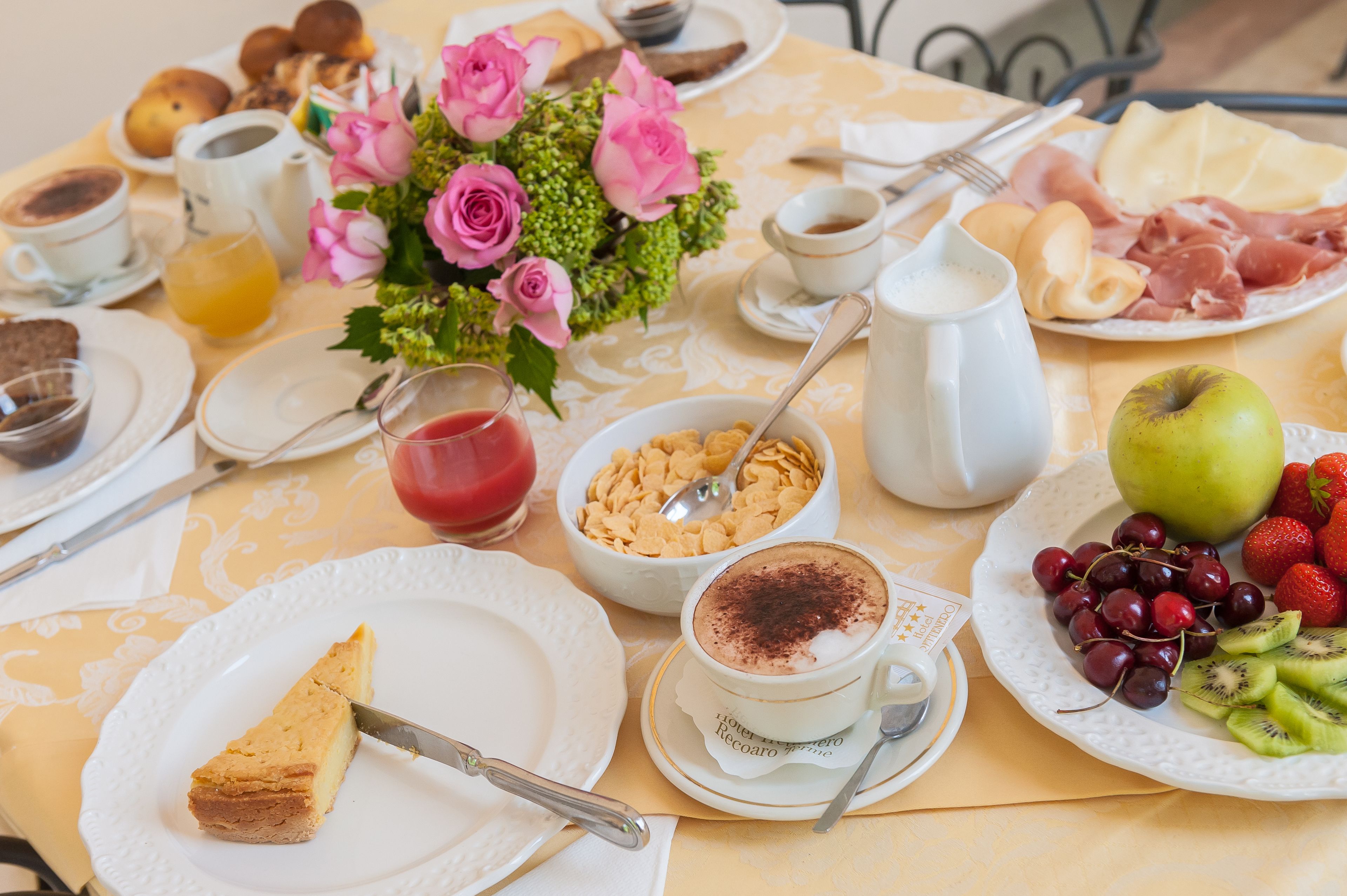 Ein Frühstückstisch mit verschiedenen Speisen, darunter ein Stück Kuchen, eine Tasse Kaffee, eine Schüssel Müsli und ein Teller Obst.